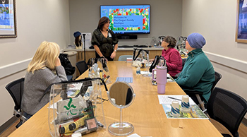 POV, looking down a conference table at a video screen showing colorful graphics. The table is in a small room with art on the left and right walls. Seated at the table are three women. Another  woman is standing to the front and left side of the screen. In the foreground on the table are standing mirrors and beauty products.