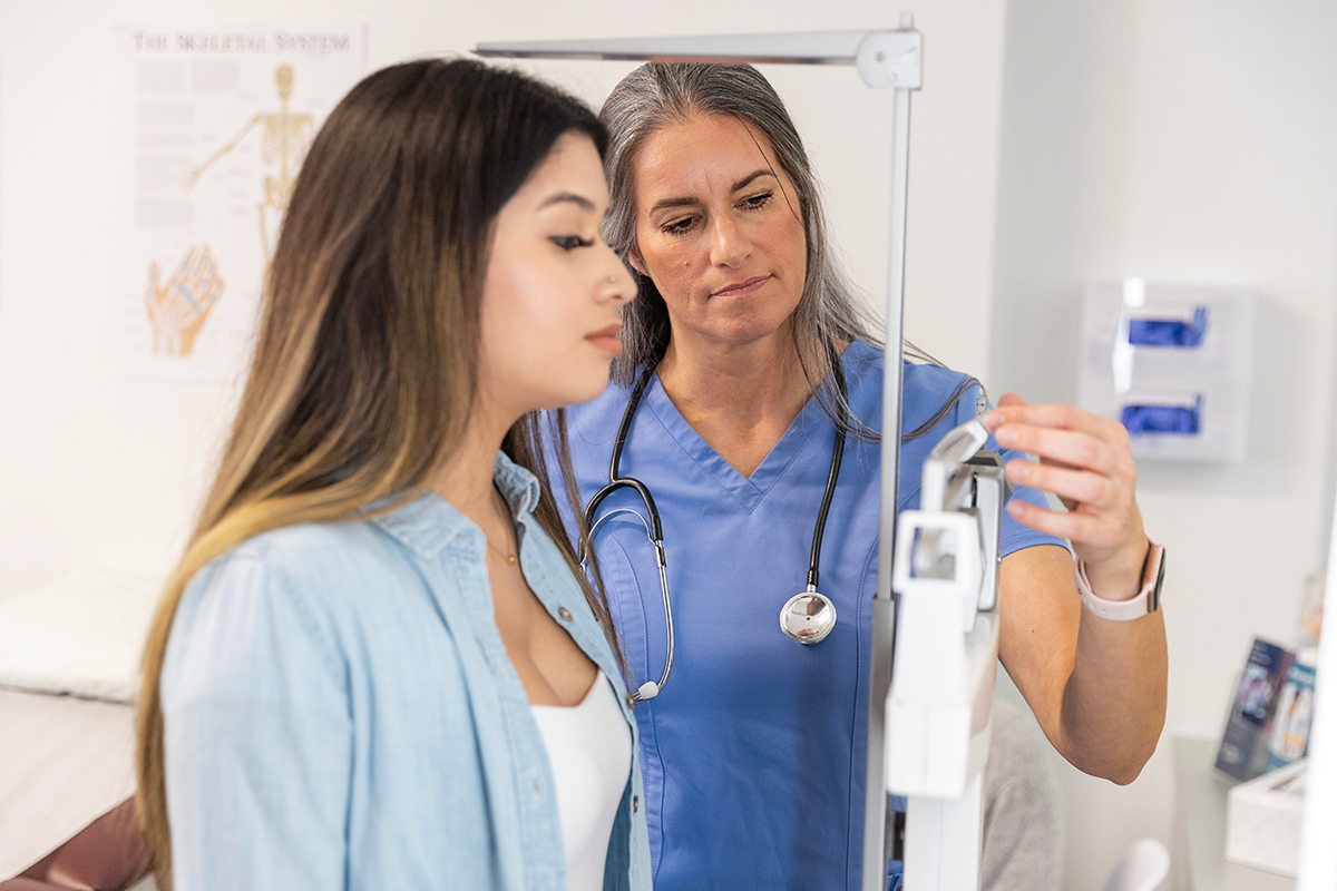 Young woman in a white top and light blue shirt looks at the reading on a scale as she is weighed by a older woman -- a nurse or doctor --  who is wearing blue scrubs and has a stethoscope around her neck..