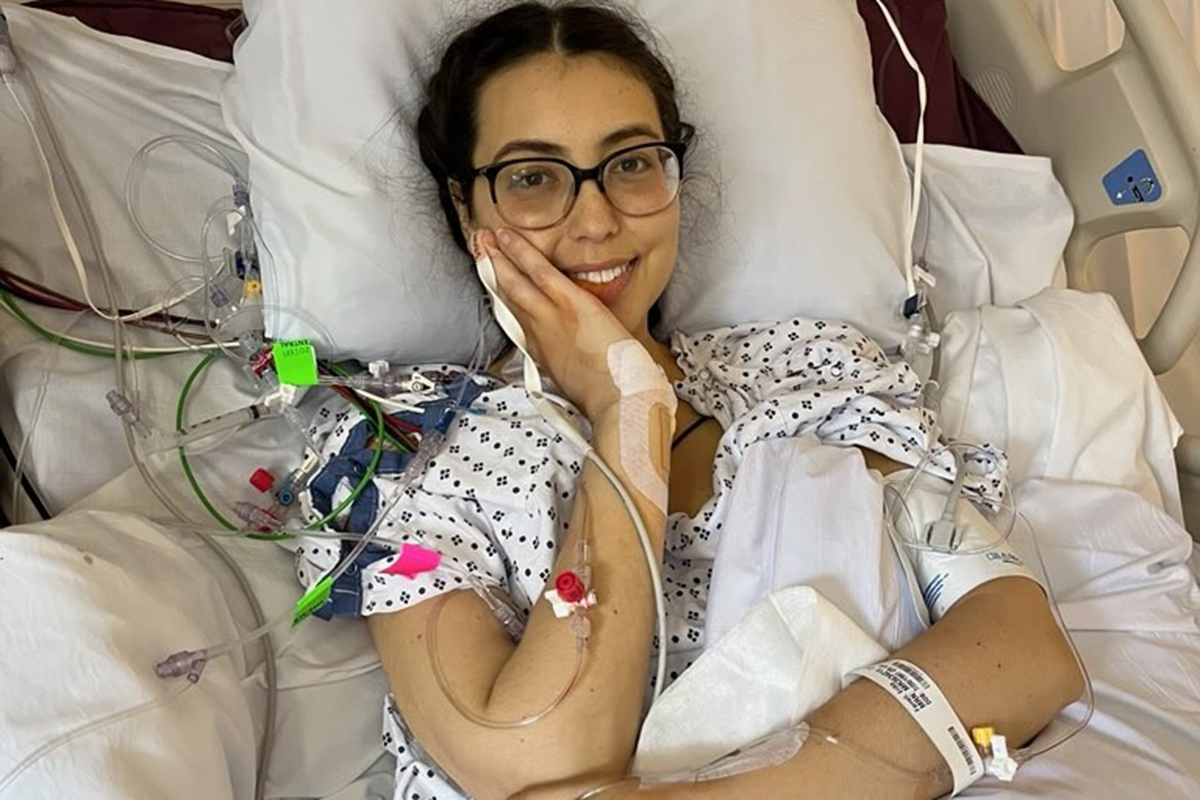 A smiling woman with dark hair and glasses sits up  in a hospital bed. She's wearing a hospital gown and has an id bracelet and IVs attateched to her arms.