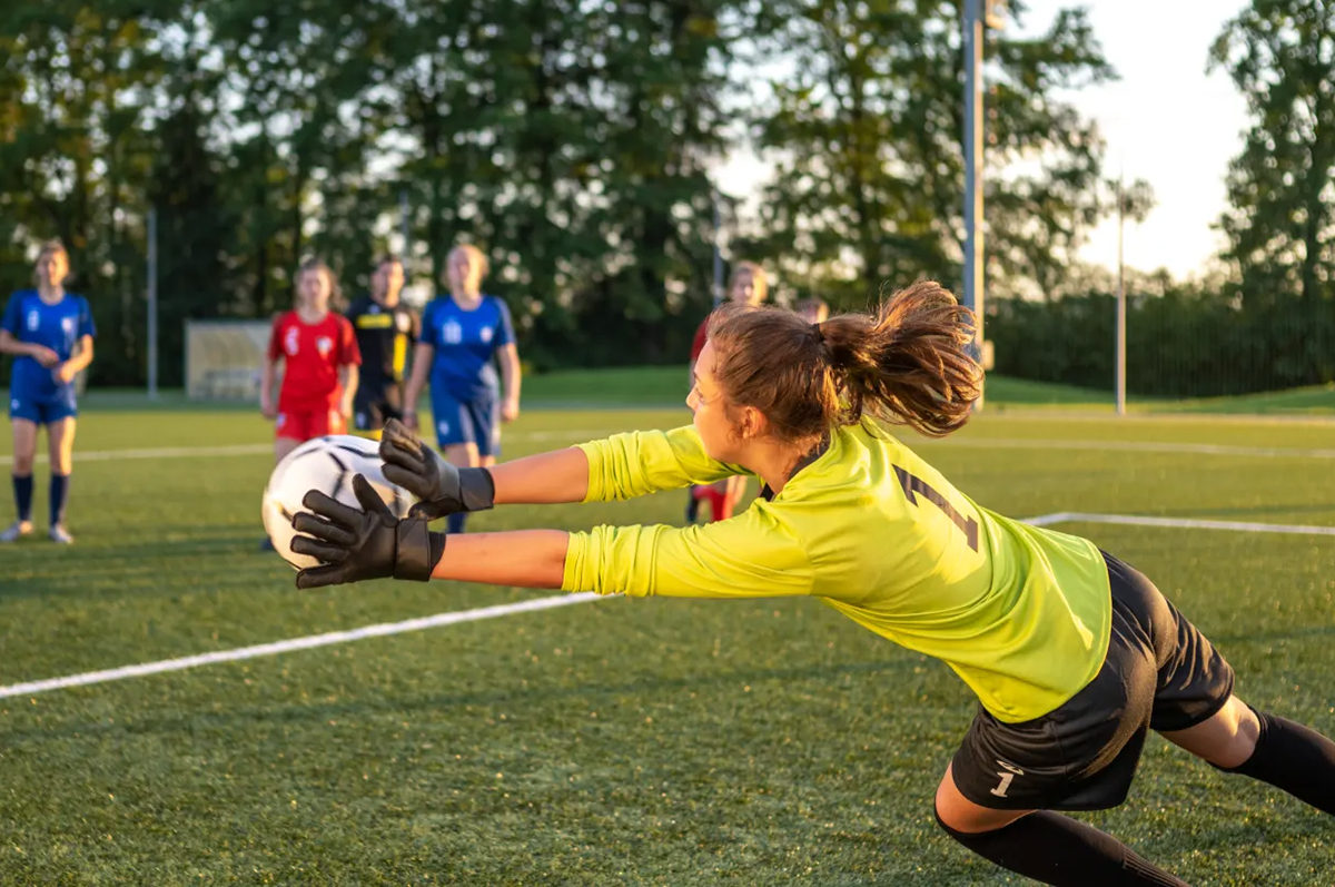 a young woman in a yellow jersey dives to catch a ball on a soccer pitch with four other players in a blurred background with trees in the distance.