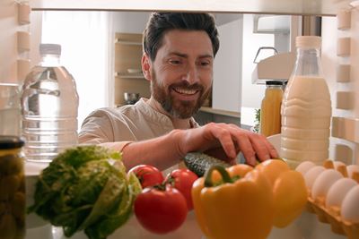 man with brown hair and trimmed beard reaches into refrigerator where there are fresh vegetables, eggs and beverages on a shelf. Photo angle is from inside  back of fridge with man facing into  it..