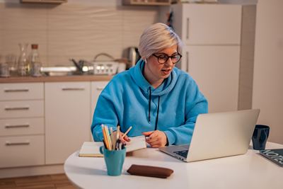 A woman with blonde hair, glasses and a light blue hooded sweatshirt sits at a table with a laptop, looking at the screen. Ont the table is a light blue cup with pencils. Setting appears to be a beige home kitchen with cabinets drawers and sink.