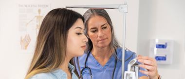 A young woman looks at the scale reading as she is weighed by an older medical professional wearing blue scrubs and stethoscope around her neck.