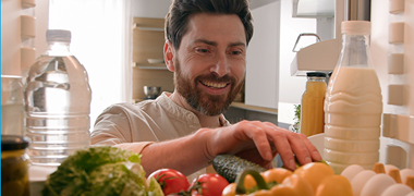 man with brown hair and trimmed beard reaches into refrigerator where there are fresh vegetables, eggs and beverages on a shelf. Photo angle is from inside  back of fridge with man facing into  it..