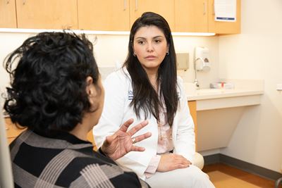 Two women, seated in a hospital exam room, face each other in discussion. In the foreground, one woman, in plaid, faces away and gestures with her left hand. The other, in a white clinical jacket, faces the camera. 